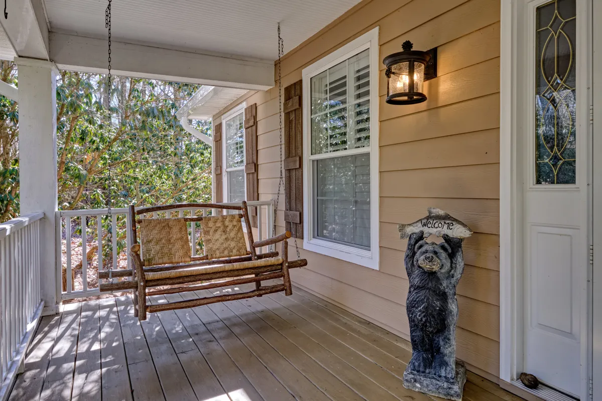 This exterior image features a cozy swinging bench on a welcoming porch, accented by a bear statue, front door, and wall lantern. The lush greenery and tranquil porch scene evoke a sense of relaxation and connection with nature, ideal for unwinding after a day of local adventures.
