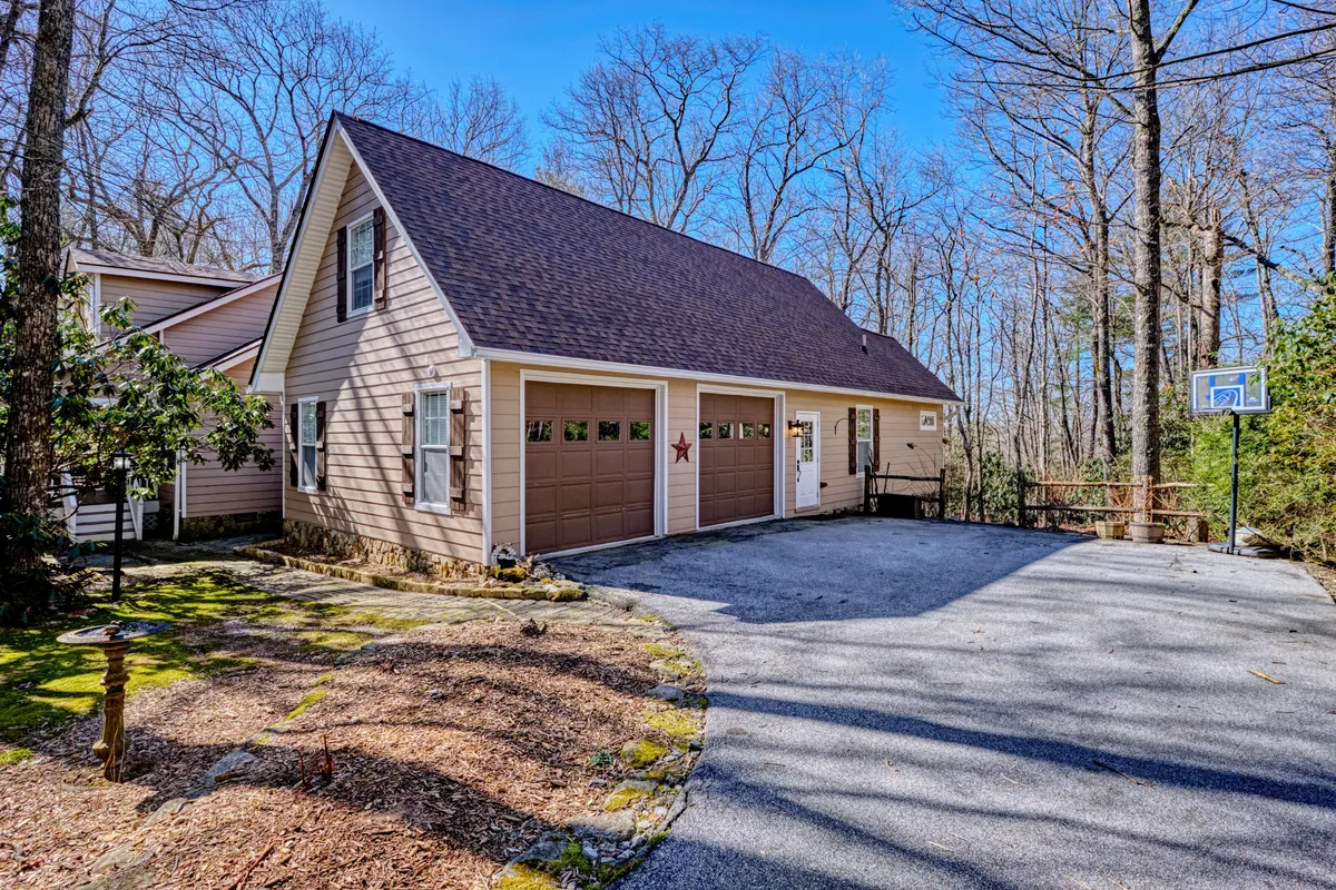 A welcoming exterior shot featuring the house facade with garage doors, a basketball hoop for recreation, and ample paved parking surrounded by trees. The scene feels open and accessible, hinting at the ease of arrival and departure in a peaceful mountain environment.
