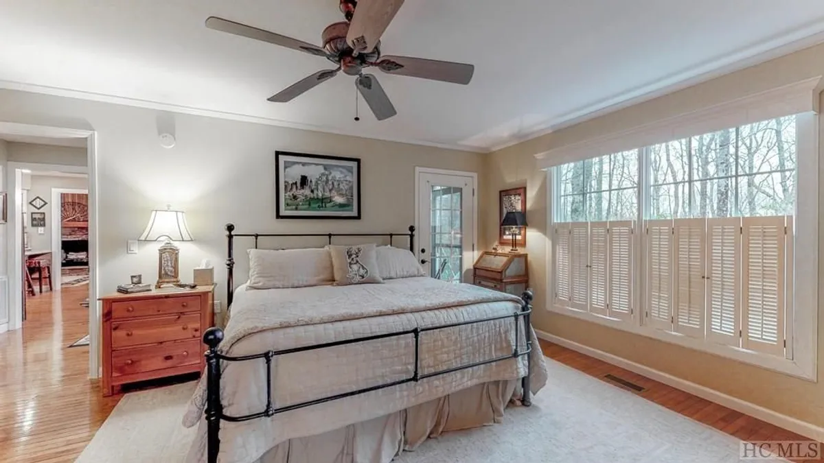 This image features a serene bedroom with a large, neatly made bed, a window with shutters allowing sunlight to gently fill the room, a nightstand with a reading lamp, and tasteful framed artwork. The setting is simple yet cozy—ideal for a queen or guest bedroom that promises comfort and privacy.