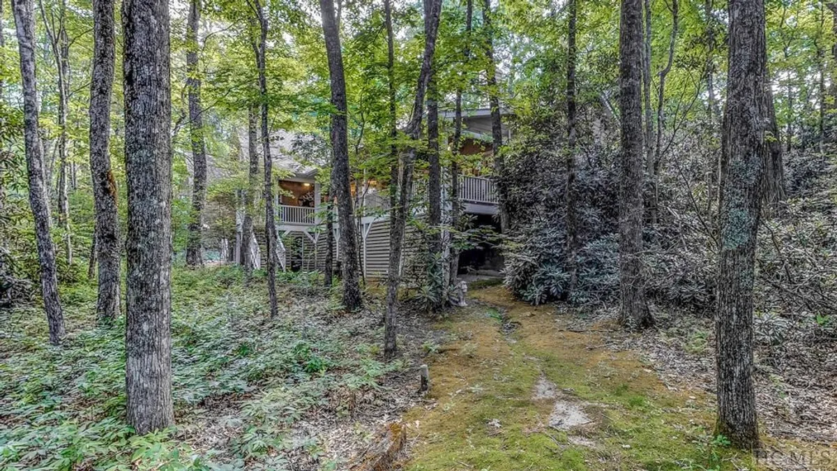 A view of the wooded property exterior with a clear pathway leading to the house, complemented by a cozy fire pit area set among mature trees. The scene emphasizes the tranquility of the natural setting and the inviting space for gatherings under the trees.