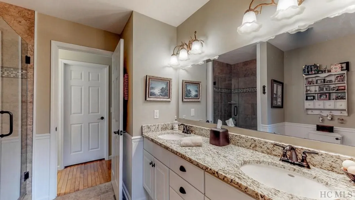 This interior image showcases a beautifully updated bathroom, featuring a sleek granite countertop with dual sinks, elegant fixtures, framed artwork, and a spacious shower area. The materials and layout evoke a sense of calm and spa-like luxury.