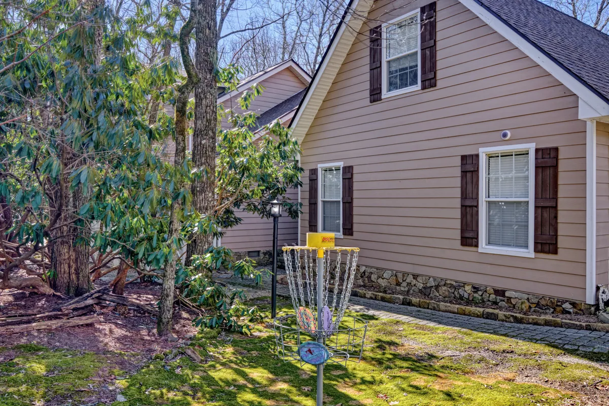 The image shows a disc golf basket set on a lush, green lawn with the house facade in the background. The surrounding landscape is vibrant and serene, suggesting ample space for outdoor activities and family play. The presence of the disc golf basket directly connects to active family fun on the property and hints at the natural beauty of the area.