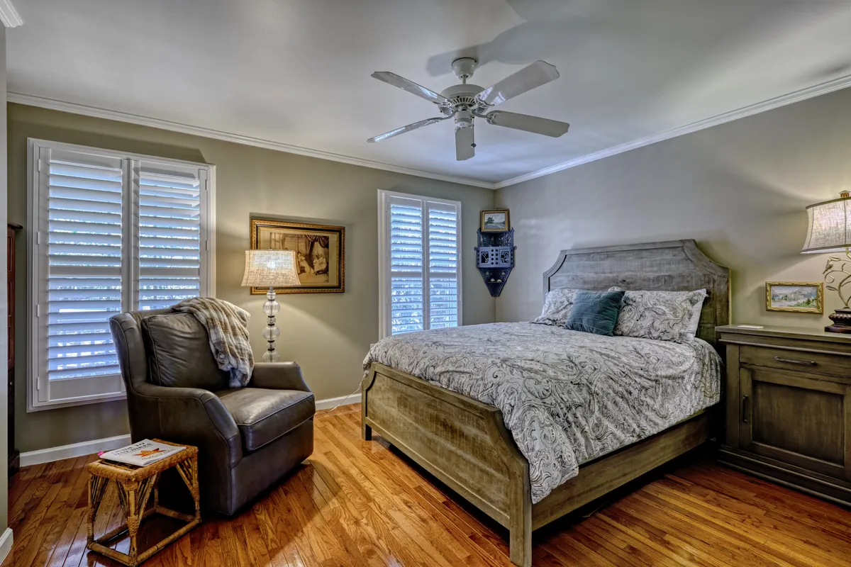 This interior image shows a cozy bedroom with a comfortable bed dressed in soft linens, an inviting armchair perfect for reading or quiet reflection, a table lamp casting a gentle glow, framed artwork on the walls, and classic shutters filtering in natural light. The scene exudes warmth and tranquility, ideal for a restful retreat.