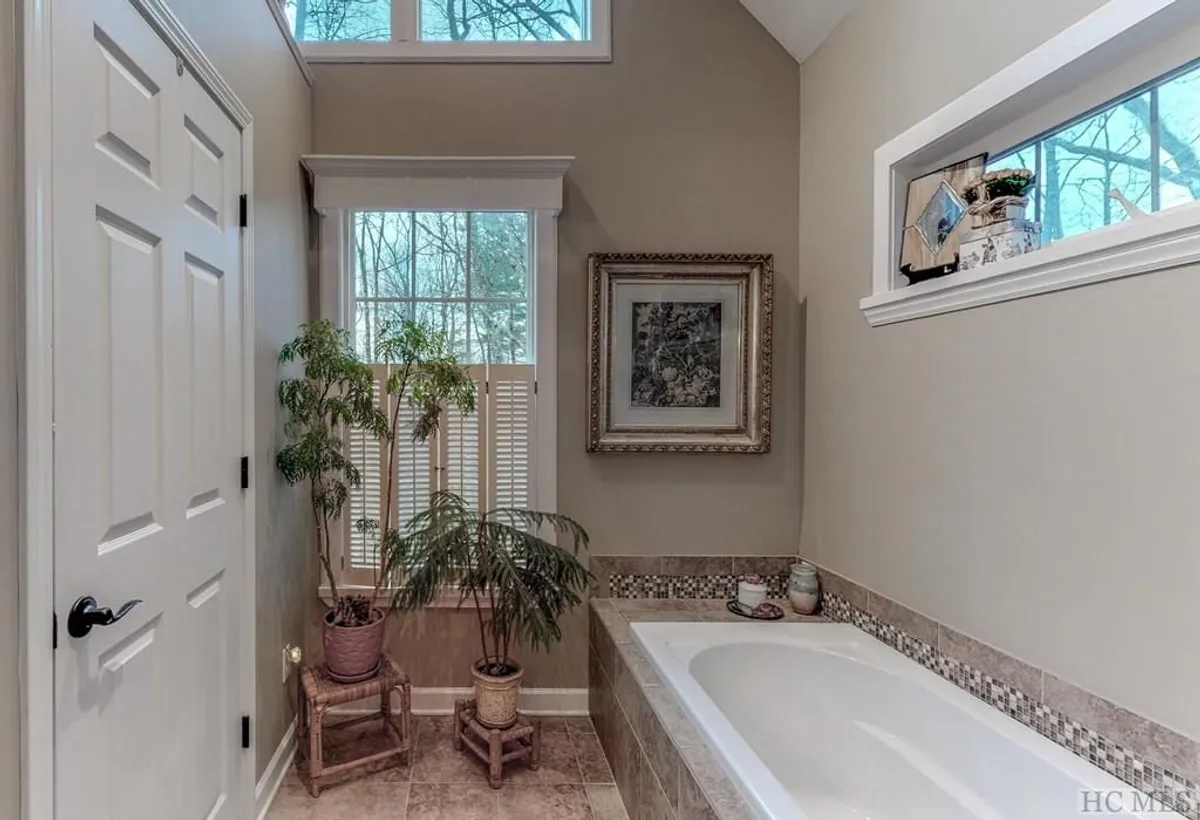 A tranquil soaking tub bathed in natural light, surrounded by lush potted plants and elegant framed artwork. The windows offer a calming view of greenery outside, suggesting a seamless connection with nature and a spa-like, meditative ambiance.
