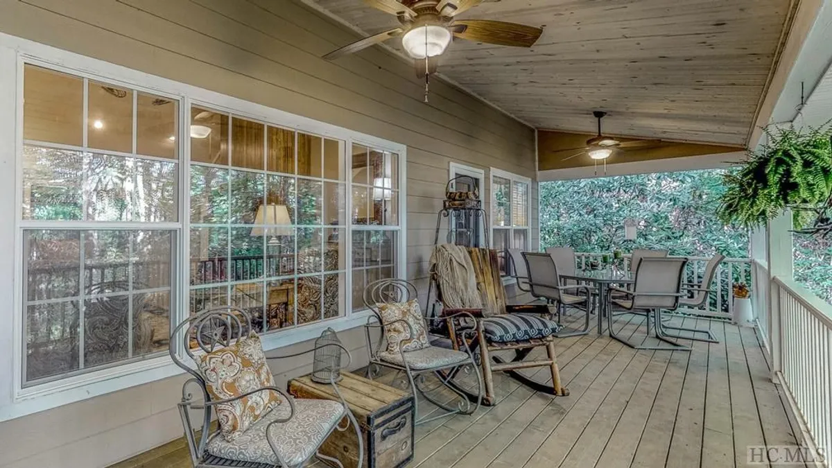 This image captures a spacious covered porch adorned with classic rocking chairs, a dining table perfect for gatherings, lush hanging ferns, and large windows that invite sunlight and forest views. The tranquil atmosphere showcases both comfort and connection with the outdoors, ideal for enjoying coffee, reading, or alfresco meals.